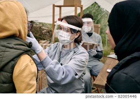 Young female medical worker in protective face shield checking throat of refugee child outdoors Young female medical worker in protective face shield checking throat of refugee child outdoors 82005446
