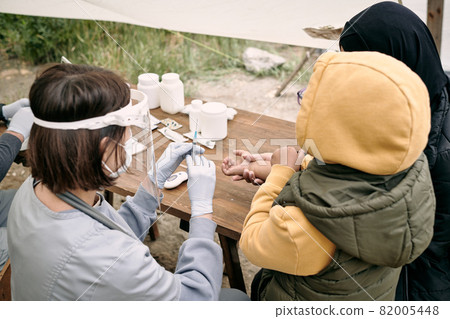 Nurse in gloves and mask preparing syringe while vaccinating refugee child outdoors 82005448
