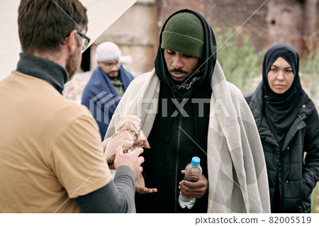 Social worker giving water and cereal to black man under plaid while providing food to refugees outdoors Social worker giving water and cereal to black man under plaid while providing food to refugees outdoors 82005519