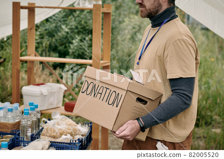 Bearded volunteer with badge carrying cardboard box with donation to table with goods for homeless Bearded volunteer with badge carrying cardboard box with donation to table with goods for homeless 82005520