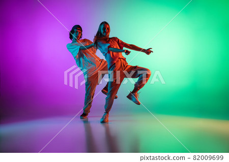 Young hip-hop dancers, stylish emotive girl and boy in action and motion in casual sports youth clothes on gradient multi colored background at dance hall in neon light. Young hip-hop dancers, stylish emotive girl and boy in action and motion in casual sports youth clothes on gradient multi colored background at dance hall in neon light. 82009699