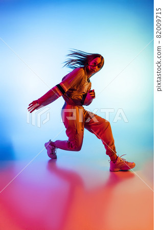 Young sportive girl dancing hip-hop in stylish clothes on colorful background at dance hall in neon light. Youth culture, movement, style and fashion, action. Young sportive girl dancing hip-hop in stylish clothes on colorful background at dance hall in neon light. Youth culture, movement, style and fashion, action. 82009715