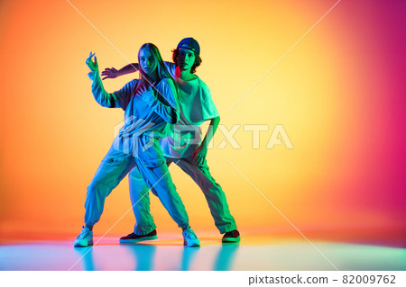 Young hip-hop dancers, stylish emotive girl and boy in action and motion in casual sports youth clothes on gradient multi colored background at dance hall in neon light. Young hip-hop dancers, stylish emotive girl and boy in action and motion in casual sports youth clothes on gradient multi colored background at dance hall in neon light. 82009762