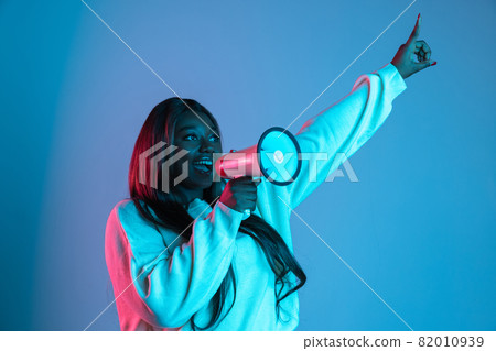 One African beautiful woman with long straight hair shouting at megaphone isolated on blue studio background in neon light. Concept of human emotions One African beautiful woman with long straight hair shouting at megaphone isolated on blue studio background in neon light. Concept of human emotions 82010939
