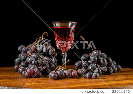 Glass with wine and dark red grapes on a black background Glass with wine and dark red grapes on a black background 82011140