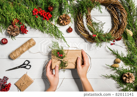 Top view of female hands wrap New Year present. Packed gifts and scrolls, spruce branches and tools on wooden table. Top view of female hands wrap New Year present. Packed gifts and scrolls, spruce branches and tools on wooden table. 82011141
