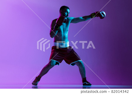 Side view full-length portrait of one professional boxer in red uniform training isolated over purple background in neon light. Straight punch move Side view full-length portrait of one professional boxer in red uniform training isolated over purple background in neon light. Straight punch move 82014342
