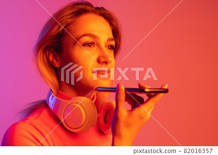 SIde view image of young beautiful girl with headphones recording voice message isolated over gradient background in neon lights. SIde view image of young beautiful girl with headphones recording voice message isolated over gradient background in neon lights. 82016357