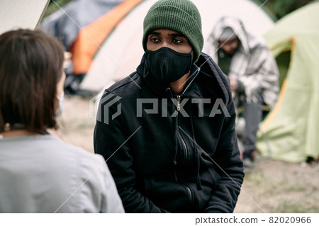Sick young Black refugee man in facial mask and hat talking to nurse in migrant camp Sick young Black refugee man in facial mask and hat talking to nurse in migrant camp 82020966
