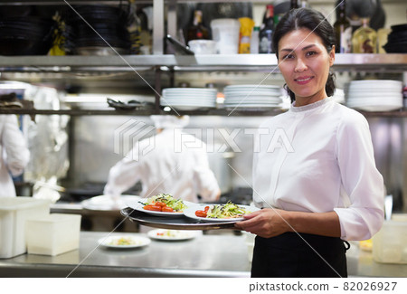 positive female waitstaff standing with tray in kitchen in restaurante 82026927