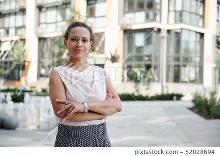 Confident business portrait of a successful middle aged business woman in casual attire posing to camera with crossed rams on the background of high buildings. Urban city backdrop Confident business portrait of a successful middle aged business woman in casual attire posing to camera with crossed rams on the background of high buildings. Urban city backdrop 82028694