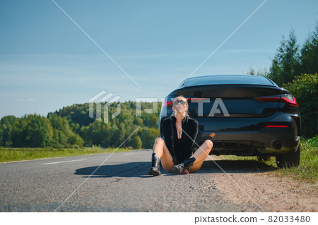 Strong woman in bodysuit and rough boots sits on asphalt by her powerful racing car 82033480