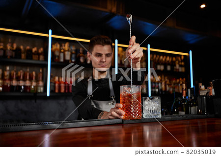 Barman stirring ice cubes in cocktail glass with spoon 82035019