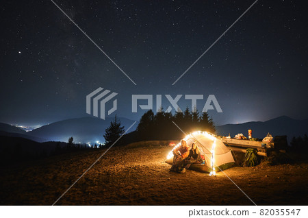 Couple travelers sitting inside illuminated camp tent and looking at evening sky with stars. Magnificent view of blue starry sky over mountain valley. Concept of travelling, hiking, night camping. Couple travelers sitting inside illuminated camp tent and looking at evening sky with stars. Magnificent view of blue starry sky over mountain valley. Concept of travelling, hiking, night camping. 82035547