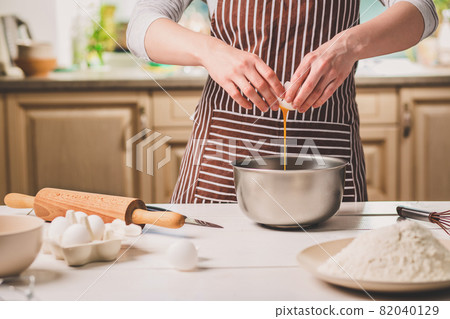 Young woman breaking egg over bowl with dough, close-up 82040129