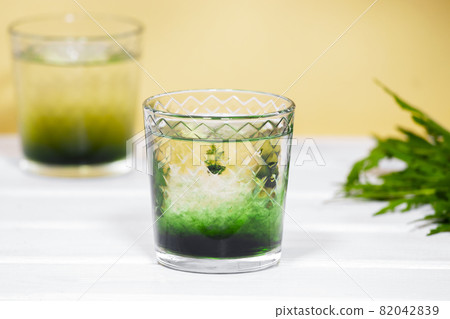 Green Chlorophyll or Chlorella drink in glass with water on white table, Orange Background. Green Drops in clear water, Antioxidant food and drinks. 82042839
