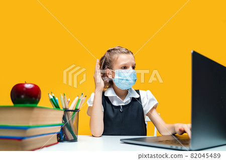 Schoolgirl raising hand and sitting at laptop wearing mask. Yellow background. Distance Learning 82045489
