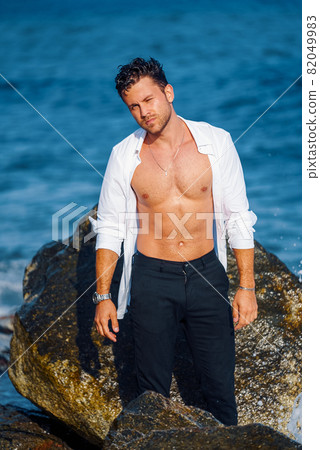 Serious confident male with wet hair and in white shirt standing near rock at seaside and looking at camera Serious confident male with wet hair and in white shirt standing near rock at seaside and looking at camera 82049983