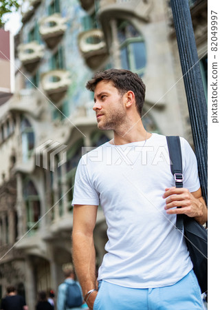 Low angle of confident young bearded male tourist in white t shirt with backpack standing with hand in pocket and looking away against old city building in summer day 82049997