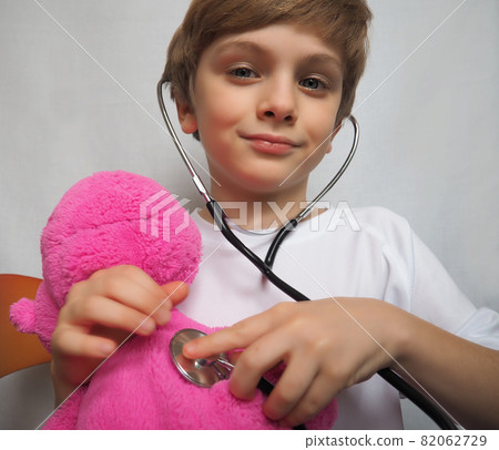 Little boy with blond hair playing with stethoscope at doctor's office. Pink plush toy in the hands of a child. Doctor game. Boy 8 years old 82062729
