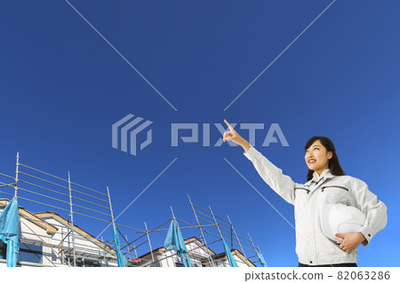 A young woman wearing work clothes, standing diagonally above and staring at the blue sky and the construction site of a detached house. A young woman wearing work clothes, standing diagonally above and staring at the blue sky and the construction site of a detached house. 82063286