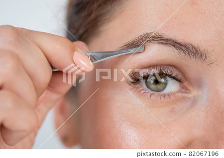 Close-up portrait of a caucasian woman doing eyebrow correction herself with tweezers Close-up portrait of a caucasian woman doing eyebrow correction herself with tweezers 82067196
