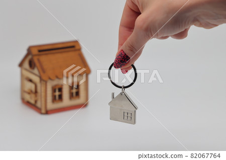 A hand holds a keychain in the shape of a house against the background of a toy deret of a withered house, mortgage, real estate purchase A hand holds a keychain in the shape of a house against the background of a toy deret of a withered house, mortgage, real estate purchase 82067764