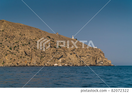 The Republic of Crimea. July 21, 2021. View from the Black Sea in warm sunny weather on the lighthouse and buildings on Mount Meganom, which is located near the city of Sudak. The Republic of Crimea. July 21, 2021. View from the Black Sea in warm sunny weather on the lighthouse and buildings on Mount Meganom, which is located near the city of Sudak. 82072322