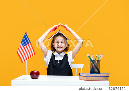 Schoolgirl in fun sitting at the desk holding book on her head. English lesson. USA flag Schoolgirl in fun sitting at the desk holding book on her head. English lesson. USA flag 82074151