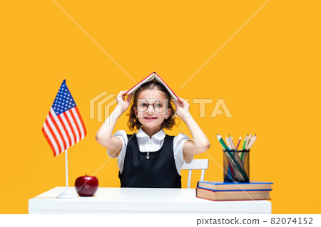 Schoolgirl in fun sitting at the desk holding book on her head. English lesson. USA flag Schoolgirl in fun sitting at the desk holding book on her head. English lesson. USA flag 82074152