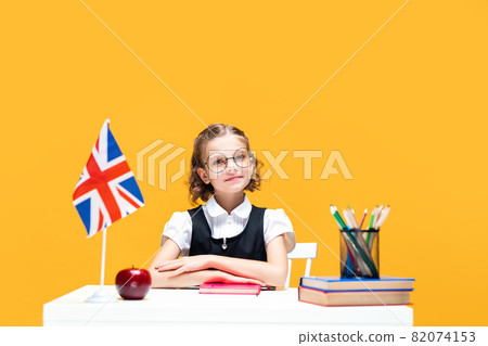 Smiling schoolgirl sitting at the desk with notebook during English lesson. Great Britain flag Smiling schoolgirl sitting at the desk with notebook during English lesson. Great Britain flag 82074153