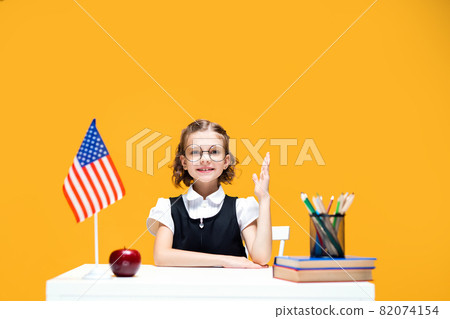 Smiling caucasian schoolgirl raising hand sitting at the desk during lesson. English lesson USA flag Smiling caucasian schoolgirl raising hand sitting at the desk during lesson. English lesson USA flag 82074154