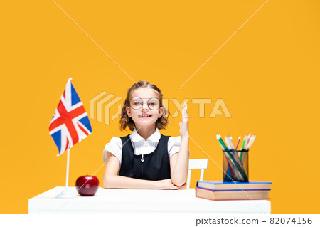 Smiling schoolgirl raising hand sitting at the desk during lesson. English lesson. Great Britain flag Smiling schoolgirl raising hand sitting at the desk during lesson. English lesson. Great Britain flag 82074156