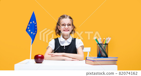 Smiling happy caucasian schoolgirl sitting at the desk during English lesson. European Union flag Smiling happy caucasian schoolgirl sitting at the desk during English lesson. European Union flag 82074162