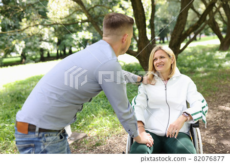 Young man provides support and walks in park with woman in wheelchair 82076877