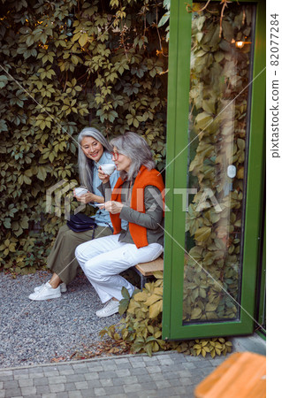 Happy mature women friends with cups of drinks sit on bench near wall with ivy 82077284