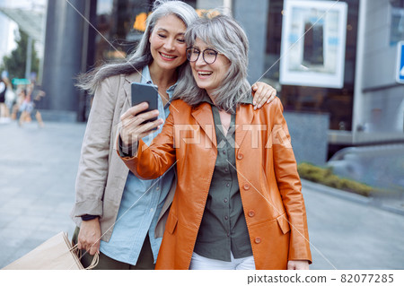 Smiling silver haired lady and companion with shopping bags take selfie on modern city street 82077285