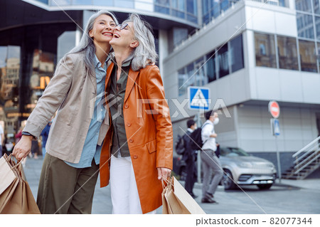 Joyful senior woman with shopping bags hug Asian lady on modern city street 82077344