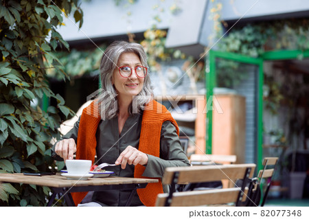 Happy mature woman with glasses eats dessert sitting at small table outdoors 82077348