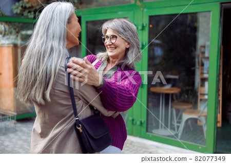 Smiling mature woman embraces female friend meeting on modern city street Smiling mature woman embraces female friend meeting on modern city street 82077349