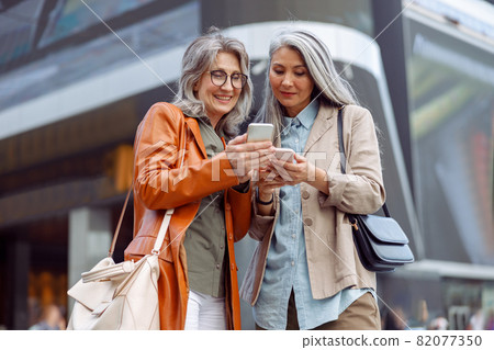 Positive grey haired ladies look at mobile phones on modern city street 82077350