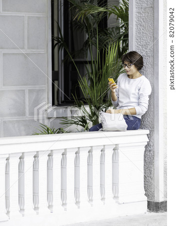 Woman texting on smartphone. Pretty female in eyeglasses sits on balcony with palm trees in flower pots. Tropical houseplant. Summer vibes. 82079042