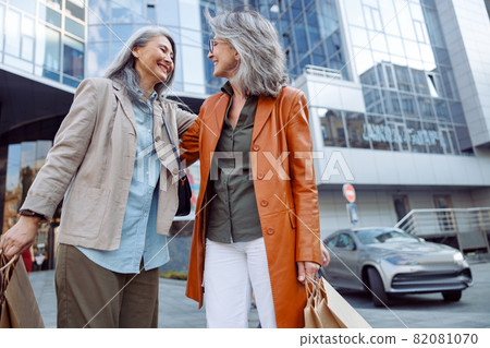 Smiling grey haired Asian woman hugs mature lady holding shopping bags on city street Smiling grey haired Asian woman hugs mature lady holding shopping bags on city street 82081070