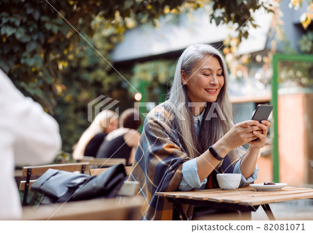 Happy mature Asian woman surfs internet on phone at small table on outdoors cafe terrace Happy mature Asian woman surfs internet on phone at small table on outdoors cafe terrace 82081071