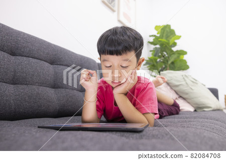 Smiling little boy looks at the touchpad lying on comfortable sofa in the living room. Enjoying playing game on digital tablet. Happy small kid using funny applications. 82084708