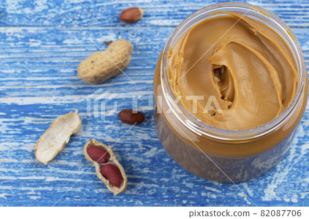 Peanut butter in an open jar and peanuts in the skin are scattered on the blue table. Peanut butter in an open jar and peanuts in the skin are scattered on the blue table. 82087706