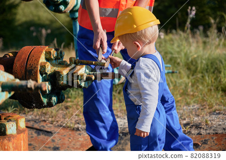 Close up view of male hands holding wrench and teaching child how to use it, while standing by rusty pipe in the fresh air. Boy in construction helmet gets acquainted with tool for nuts. Close up view of male hands holding wrench and teaching child how to use it, while standing by rusty pipe in the fresh air. Boy in construction helmet gets acquainted with tool for nuts. 82088319
