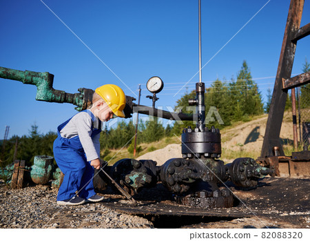 Side view of kid boy playing with spanner at pipeline. Little mechanic in construction helmet holding wrench near oil transport system for petroleum pumping station. Side view of kid boy playing with spanner at pipeline. Little mechanic in construction helmet holding wrench near oil transport system for petroleum pumping station. 82088320