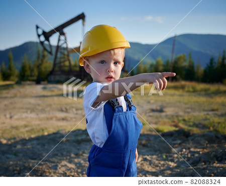 Cute child oil well worker looking at camera and pointing finger at something while standing in oil field. Adorable child wearing blue work overalls and construction helmet. 82088324