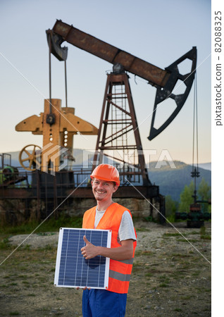 Half length portrait of cheerful serviceman in uniform holding solar battery and showing OK during shift job at oil station, concept of alternative green energy for extracting with sun battery Half length portrait of cheerful serviceman in uniform holding solar battery and showing OK during shift job at oil station, concept of alternative green energy for extracting with sun battery 82088325